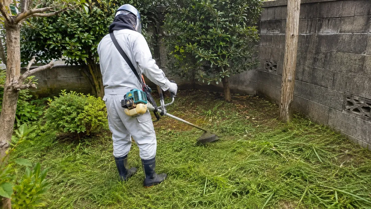 刈払機で空き家の庭の草を刈る作業員